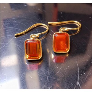 May include: A pair of gold-toned earrings featuring rectangular, faceted orange gemstones set in metal frames. Each earring is suspended from a curved gold-toned hook wire, showcasing a clear, vibrant orange hue and a polished, reflective finish.