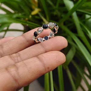 Può includere: Un anello in argento con un motivo ripetitivo di pietre rotonde nere alternate a pietre cabochon traslucide verde pallido, incastonate in una fascia continua. L'anello in metallo ha una finitura lucida con una serie di incastonature circolari.