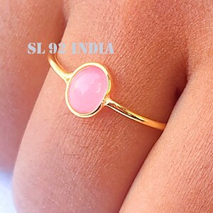 K&ouml;nnte beinhalten: Ein goldener Ring mit einem rosafarbenen ovalen Edelstein. Der Ring befindet sich an einem Finger.