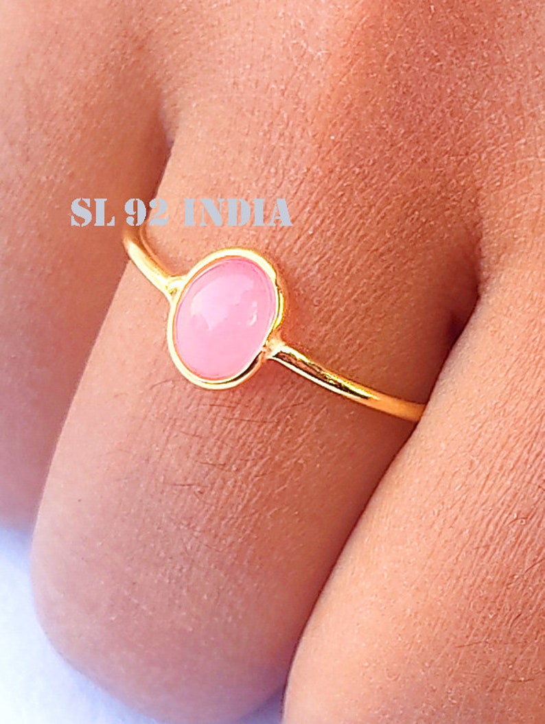 K&ouml;nnte beinhalten: Ein goldener Ring mit einem rosafarbenen ovalen Edelstein. Der Ring befindet sich an einem Finger.