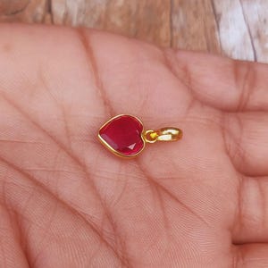 May include: A small heart-shaped pendant featuring a faceted deep red gemstone set in a polished gold-toned metal frame. The pendant includes a small circular gold-toned bail at the top for attachment to a chain or cord, showcasing a simple design.