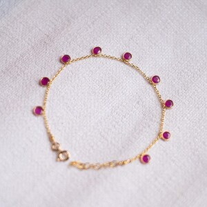 Puede incluir: Una delicada pulsera de oro con una serie de vibrantes piedras preciosas de rubí redondas. La cadena de la pulsera es de un fino eslabón dorado, y el cierre es visible. Las piedras preciosas están espaciadas uniformemente alrededor de la circunferencia de la pulsera.