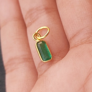 Könnte beinhalten: Ein goldener Anhänger mit einem grünen Smaragd-Edelstein. Der Anhänger ist rechteckig und hat oben eine kleine Öse zum Anbringen an einer Kette.