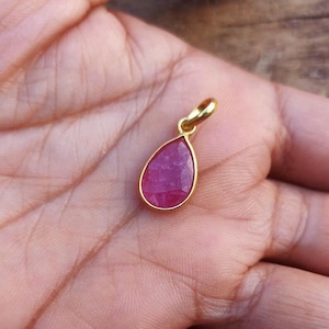 May include: A teardrop-shaped pendant with a vibrant red gemstone, set in a gold-colored frame. The pendant has a small loop at the top for a chain. The gemstone is a deep, rich red color.