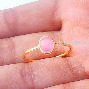 K&ouml;nnte beinhalten: Ein goldener Ring mit einem rosafarbenen ovalen Edelstein. Der Ring befindet sich an einem Finger.