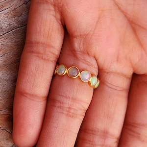 Puede incluir: Un anillo de oro con una serie de piedras de ópalo ovaladas. Los ópalos muestran una gama de colores, incluyendo blanco, rosa y verde. El anillo se muestra sobre un fondo de madera.
