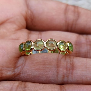 Op de afbeelding: Een gouden ring met een reeks ovale edelstenen in groen- en geeltinten. De ring heeft een gouden band, met de edelstenen gelijkmatig verdeeld over de omtrek. De ring wordt op een hand getoond.