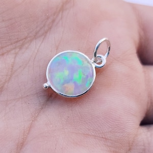 Puede incluir: Un colgante de plata con una piedra de ópalo redonda. El ópalo es iridiscente con tonos de verde, azul y rosa.