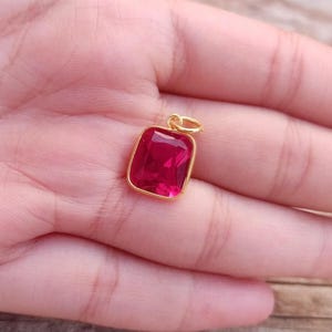 May include: A gold pendant with a rectangular, deep red gemstone. The gemstone is set in a gold frame and has a small gold loop at the top for a chain. The pendant is held in a hand.
