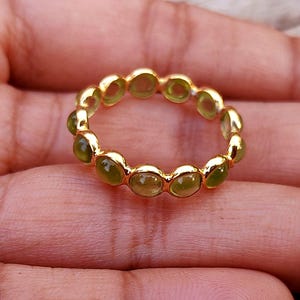 K&ouml;nnte beinhalten: Ein goldener Ring mit einem durchgehenden Band aus ovalen, hellgr&uuml;nen Edelsteinen. Der Ring ist in einem goldenen Band gefasst, und die Edelsteine sind gleichm&auml;&szlig;ig verteilt. Der Ring wird auf einer Hand gezeigt.