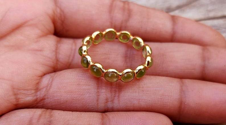 K&ouml;nnte beinhalten: Ein goldener Ring mit einem durchgehenden Band aus ovalen, hellgr&uuml;nen Edelsteinen. Der Ring ist in einem goldenen Band gefasst, und die Edelsteine sind gleichm&auml;&szlig;ig verteilt. Der Ring wird auf einer Hand gezeigt.