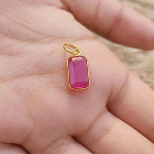 Peut inclure: Un pendentif doré avec une pierre précieuse rectangulaire de couleur magenta. La pierre est sertie dans une lunette dorée et possède une petite boucle dorée pour une chaîne. Le pendentif est tenu dans une main.