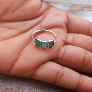 May include: A silver ring featuring three rectangular, green, translucent stones set horizontally in a row. The band is thin and metallic, showcasing a simple design that highlights the three distinct green stones centered on the finger.