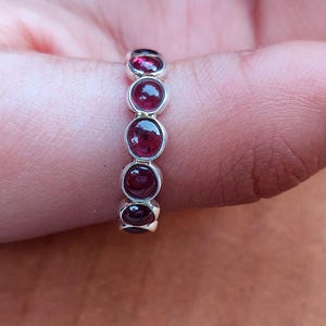 May include: A silver ring with five oval, deep red gemstones. The gemstones are set in a row along the ring's band. The ring is shown on a person's finger, with the skin tone visible in the background.