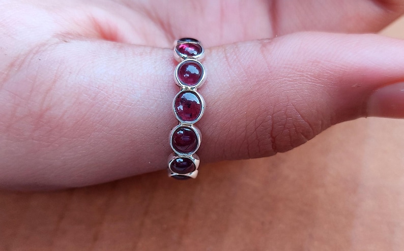 May include: A silver ring with five oval, deep red gemstones. The gemstones are set in a row along the ring's band. The ring is shown on a person's finger, with the skin tone visible in the background.