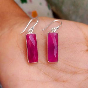 May include: A pair of drop earrings featuring rectangular, faceted fuchsia-colored gemstones set in silver-toned metal frames. Each earring hangs from a simple silver-toned hook wire, showcasing a polished finish and a vibrant, deep pink hue.