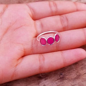 Peut inclure: Une bague en argent ornée de trois petites pierres ovales d'un rose vif disposées en ligne. L'anneau est fin et poli, présentant une finition métallique qui contraste avec la couleur vive et saturée des trois pierres circulaires incrustées.