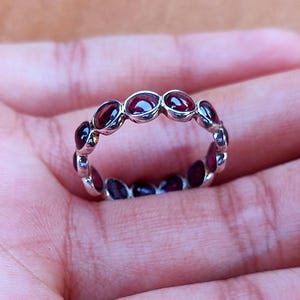 May include: A silver ring featuring a series of oval-cut, deep red gemstones. The gemstones are set closely together, forming a continuous band around the ring. The ring is displayed on a hand, showcasing its design and color.