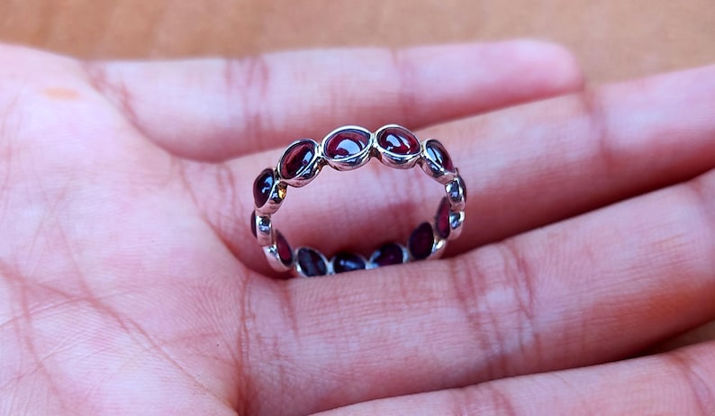 May include: A silver ring featuring a series of oval-cut, deep red gemstones. The gemstones are set closely together, forming a continuous band around the ring. The ring is displayed on a hand, showcasing its design and color.