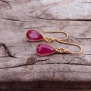Peut inclure: Une paire de boucles d'oreilles pendantes dorées avec des pierres précieuses rouges en forme de goutte. Les boucles d'oreilles sont présentées sur une surface en bois vieilli, soulignant le contraste entre le métal poli et le fond rustique. Bijoux élégants.