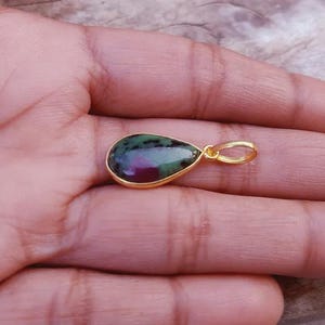 May include: A teardrop-shaped pendant with a gold-colored setting. The gemstone features a mix of green and red hues. The pendant is displayed on a hand, showcasing the jewelry's design and color.