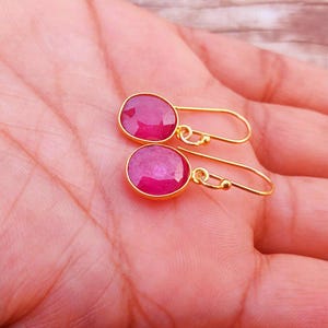 May include: A pair of gold-toned drop earrings featuring round, faceted, deep pink ruby-coloured stones set in smooth metal bezels. The earrings are attached to simple, curved gold-toned wire hooks that allow for movement and dangle from the earlobe.