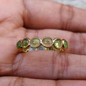 K&ouml;nnte beinhalten: Ein goldener Ring mit einer Reihe runder, hellgr&uuml;ner Edelsteine. Der Ring ist in einem goldenen Band gefasst, wobei die Edelsteine gleichm&auml;&szlig;ig &uuml;ber die Oberseite verteilt sind. Der Ring wird auf einer Hand gezeigt.