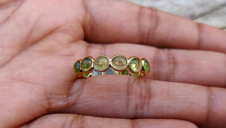 K&ouml;nnte beinhalten: Ein goldener Ring mit einer Reihe runder, hellgr&uuml;ner Edelsteine. Der Ring ist in einem goldenen Band gefasst, wobei die Edelsteine gleichm&auml;&szlig;ig &uuml;ber die Oberseite verteilt sind. Der Ring wird auf einer Hand gezeigt.