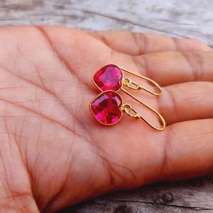 May include: A pair of gold-toned hook earrings featuring faceted, heart-shaped, vibrant pink-red stones. The earrings have a simple wire hook design that allows the heart-shaped pendants to dangle freely from the base of the metal hook.
