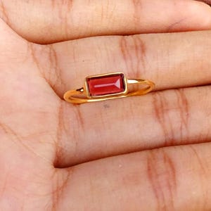 May include: A gold ring featuring a single rectangular baguette-cut deep red garnet gemstone set horizontally in a simple polished gold band. The ring has a minimalist design with a smooth, thin metal shank that encircles the finger.
