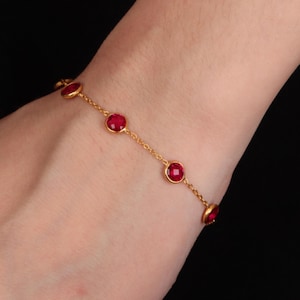 Peut inclure: Un bracelet doré avec une fine chaîne et une série de pierres précieuses rouges rondes. Les pierres sont espacées le long de la chaîne, créant un design simple et élégant. Le bracelet est porté sur un poignet.