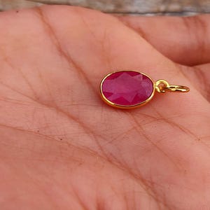 May include: A gold pendant featuring a faceted, oval-shaped, deep red gemstone. The gemstone is set in a gold bezel and has a small gold loop for attaching to a chain. The pendant is shown against a skin-toned background.