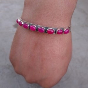 May include: A silver bangle bracelet with pink oval-shaped gemstones. The bracelet is on a person's wrist.