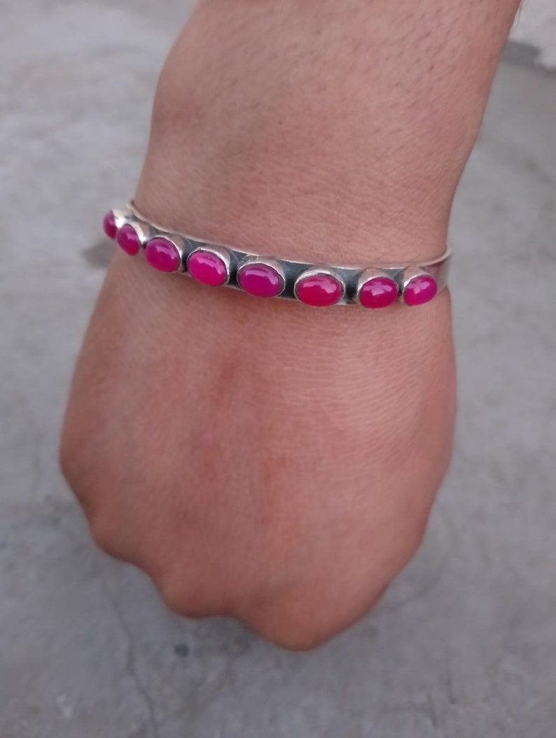 May include: A silver bangle bracelet with pink oval-shaped gemstones. The bracelet is on a person's wrist.