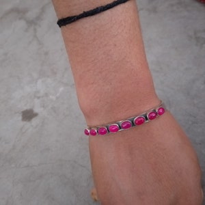 May include: A silver bangle bracelet with pink oval-shaped stones set in the metal. The bracelet is on a person's wrist.