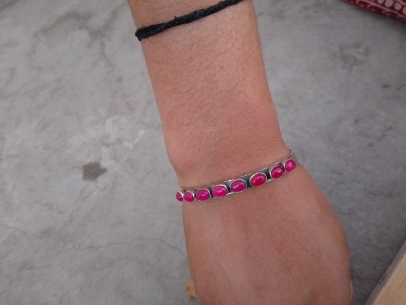 May include: A silver bangle bracelet with pink oval-shaped stones set in the metal. The bracelet is on a person's wrist.