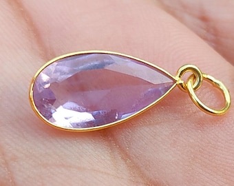 Puede incluir: Un colgante en forma de lágrima con una piedra preciosa de color púrpura claro engastada en un bisel dorado. El colgante tiene un pequeño lazo en la parte superior para una cadena. La piedra está facetada. Joyería.