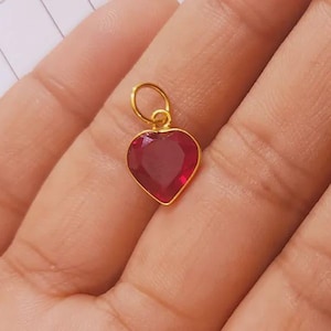 May include: A heart-shaped pendant featuring a deep red faceted gemstone set in a thin gold-toned metal frame. The pendant includes a small circular gold-toned jump ring at the top for attachment to a chain or cord, showcasing a polished finish.