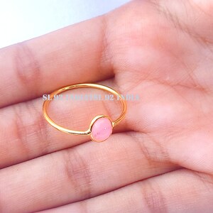 K&ouml;nnte beinhalten: Ein goldener Ring mit einem rosafarbenen ovalen Edelstein. Der Ring befindet sich an der Hand einer Person.
