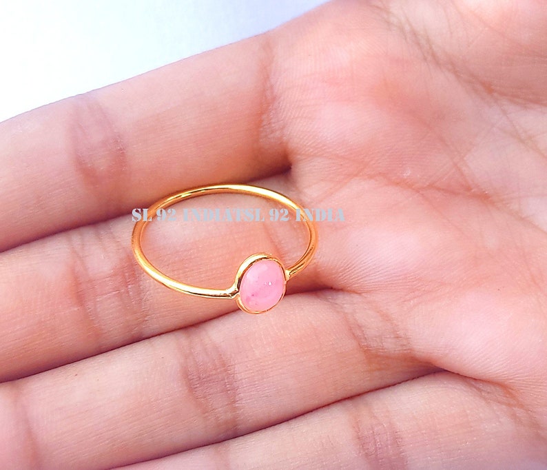 K&ouml;nnte beinhalten: Ein goldener Ring mit einem rosafarbenen ovalen Edelstein. Der Ring befindet sich an der Hand einer Person.