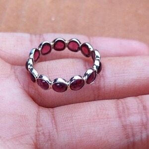 May include: A silver ring with a continuous band of oval-shaped, deep red gemstones. The ring is set against a neutral background, showcasing the jewelry's design and color. The gemstones are evenly spaced around the ring's circumference.