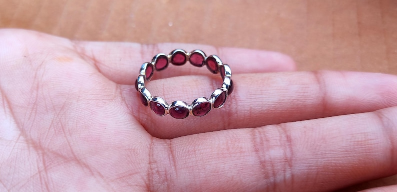 May include: A silver ring with a continuous band of oval-shaped, deep red gemstones. The ring is set against a neutral background, showcasing the jewelry's design and color. The gemstones are evenly spaced around the ring's circumference.