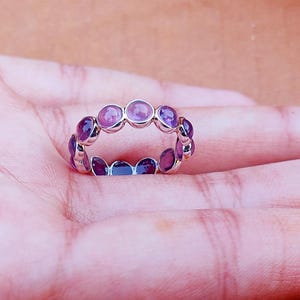 May include: A silver ring featuring a continuous band of alternating round, purple gemstones. The ring is set against a light pink background. The gemstones are a mix of light and dark purple hues.