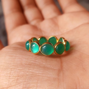 May include: A gold band ring featuring a series of round, smooth, translucent green cabochon stones set side-by-side in a continuous circular arrangement. The ring has a polished metallic finish and a vibrant, consistent green colour throughout.