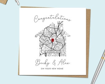 May include: A white card with black text that reads "Congratulations Becky & Alex on your new home". The card features a black and white illustration of a house with a red pin in the center. The house is made up of a map of a city with a street sign that reads "4 Riverside Drive".