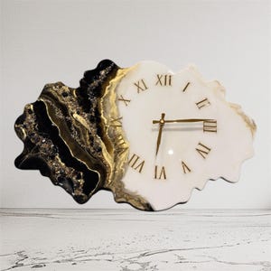 May include: A unique wall clock with a white face and gold Roman numerals. The left side features a black and gold marbled design. The clock hands are gold. The clock has a freeform shape, adding a modern touch to any space.