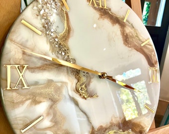 May include: Round wall clock with a white and brown marbled design, gold Roman numerals, and gold hands. The clock face has a glossy finish and is accented with gold and crystal embellishments. The clock is displayed on a wooden easel.
