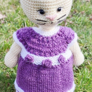 May include: A crocheted cat doll wearing a purple dress with white trim and white crocheted shoes. The doll has a white face with black eyes and a black nose.