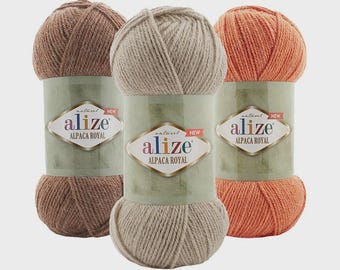 Puede incluir: Tres ovillos de hilo Alize Alpaca Royal en tonos marrón, beige y naranja. Cada ovillo está envuelto con una etiqueta verde con el nombre de la marca y los detalles del producto. El hilo está bien enrollado, mostrando su textura.