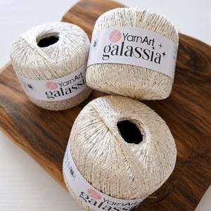 Può includere: Tre gomitoli di filato YarnArt Galassia, ciascuno caratterizzato da un filo metallico color crema intrecciato con riflessi argentati scintillanti. Ogni gomitolo è avvolto da un'etichetta di carta bianca con il marchio YarnArt e Galassia.